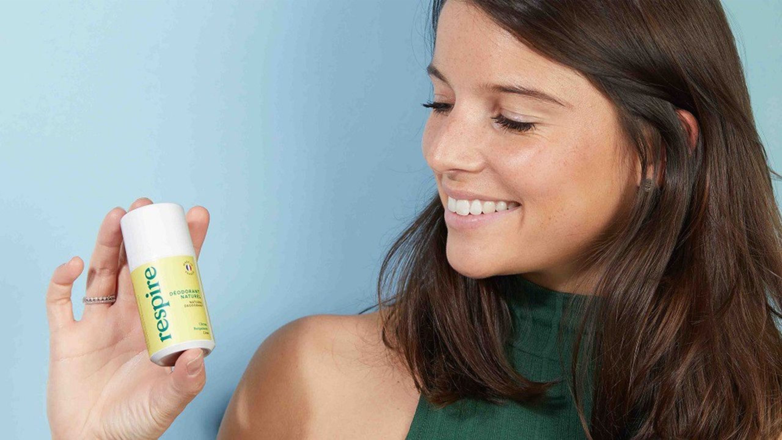 justine hutteau devant un fond bleu portant dans sa main un deodorant respire en stick au thé blanc
